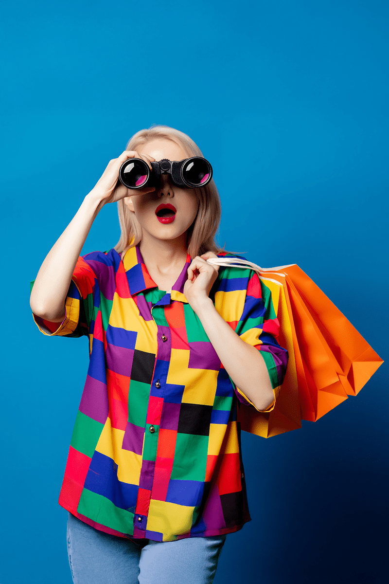 woman-searching-with-binoculars-and-shopping-bags Blonde woman wearing a colorful shirt holding orange shopping bags and looking through binoculars against a blue background, expressing search and discovery.